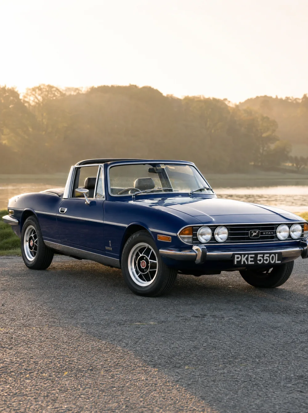 Klassieke Triumph Stag grand tourer uit 1973 in saffierblauw, driekwart vooraanzicht in gouden ochtendlicht.